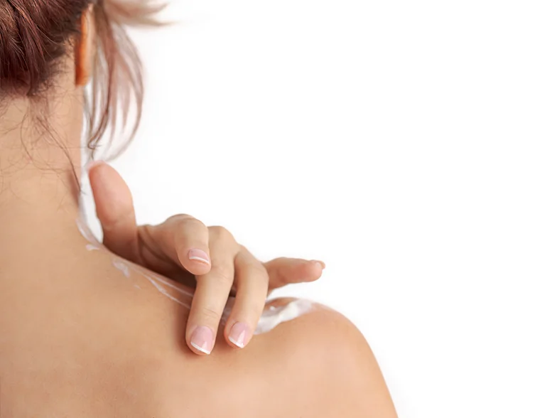 A close-up of a woman's shoulder and neck, as she gently applies cream on her skin.